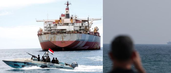 A large cargo ship at sea with a smaller military boat in the foreground, and a blurred person looking out to sea on the right.